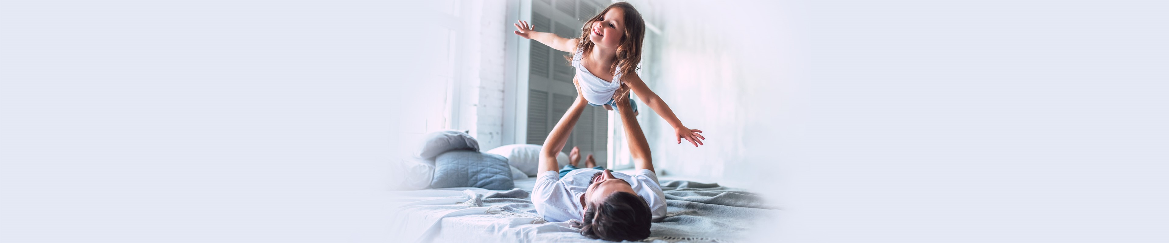 Egy fekvő férfi látható a képen, aki a kislányát tartja a magasba, aki széttárt karokkal úgy tesz, mintha repülne.